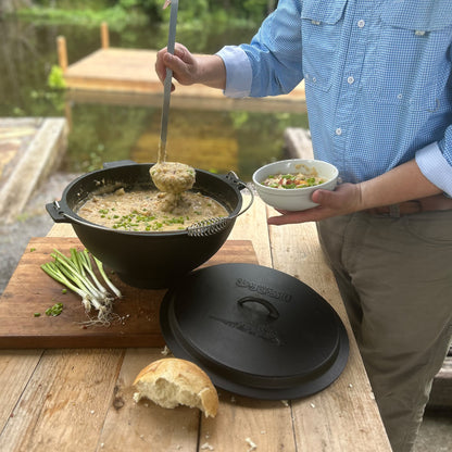 Person serving chowder from a large cast iron pot on a wooden table outdoors.