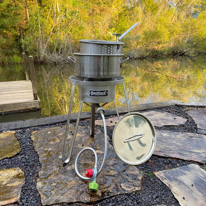 Outdoor stainless cooker with stainless fry pot sitting on a patio setting by water with autumn trees in the background.