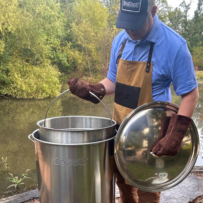 Person holding a stainless steel stockpot with basket and lid outdoors by a body of water