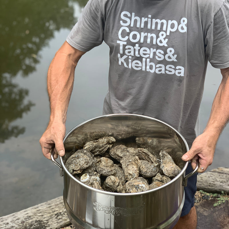 A man wearing a  gray t-shirt with white text stands by a body of water holding a large stainless steel pot filled with oysters. The t-shirt reads 'Shrimp & Corn & Taters & Kielbasa'.