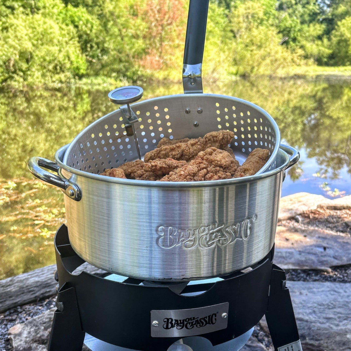 Fry pot with fried chicken on an outdoor cooker ouside by a lake.