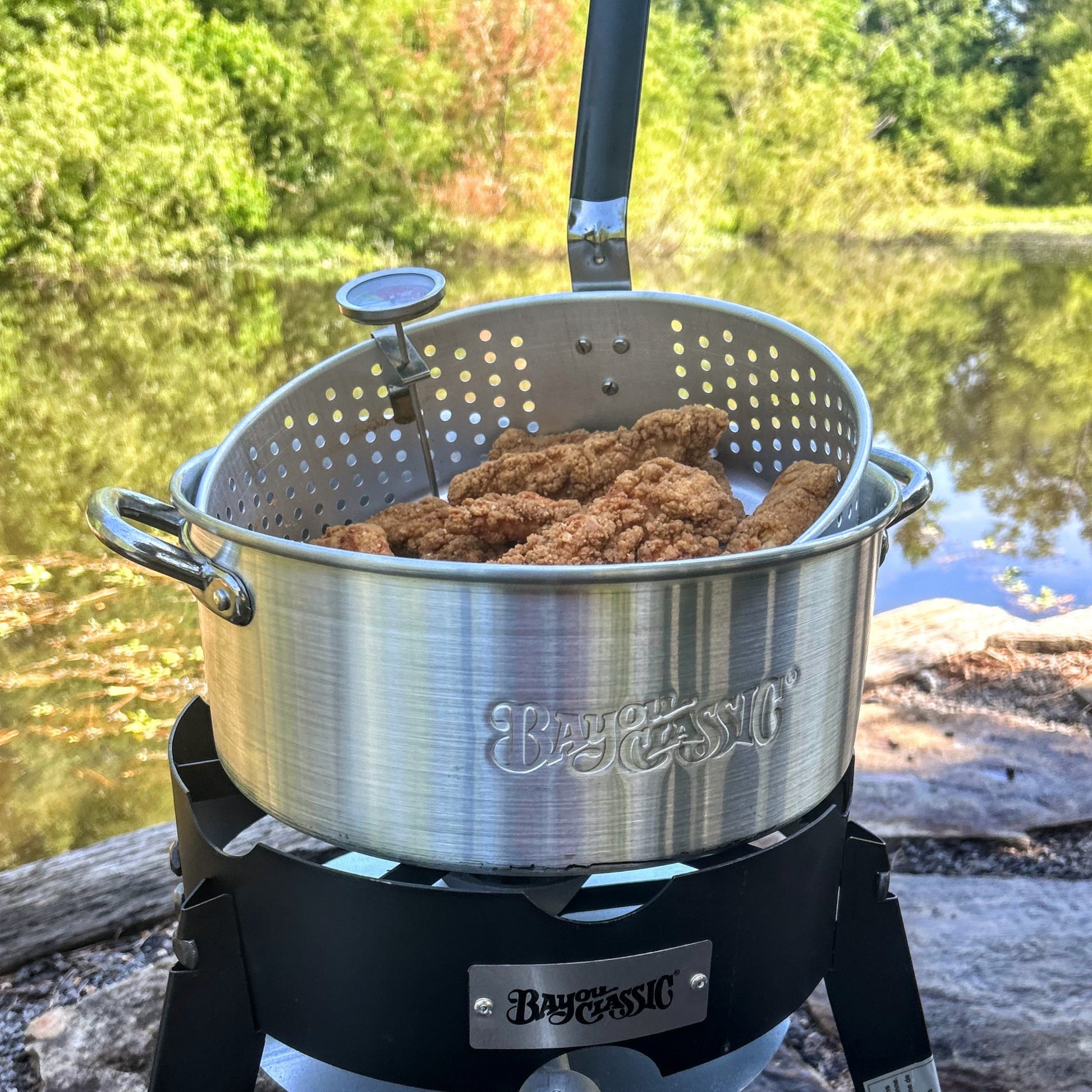 Fry pot with fried chicken on an outdoor cooker ouside by a lake.