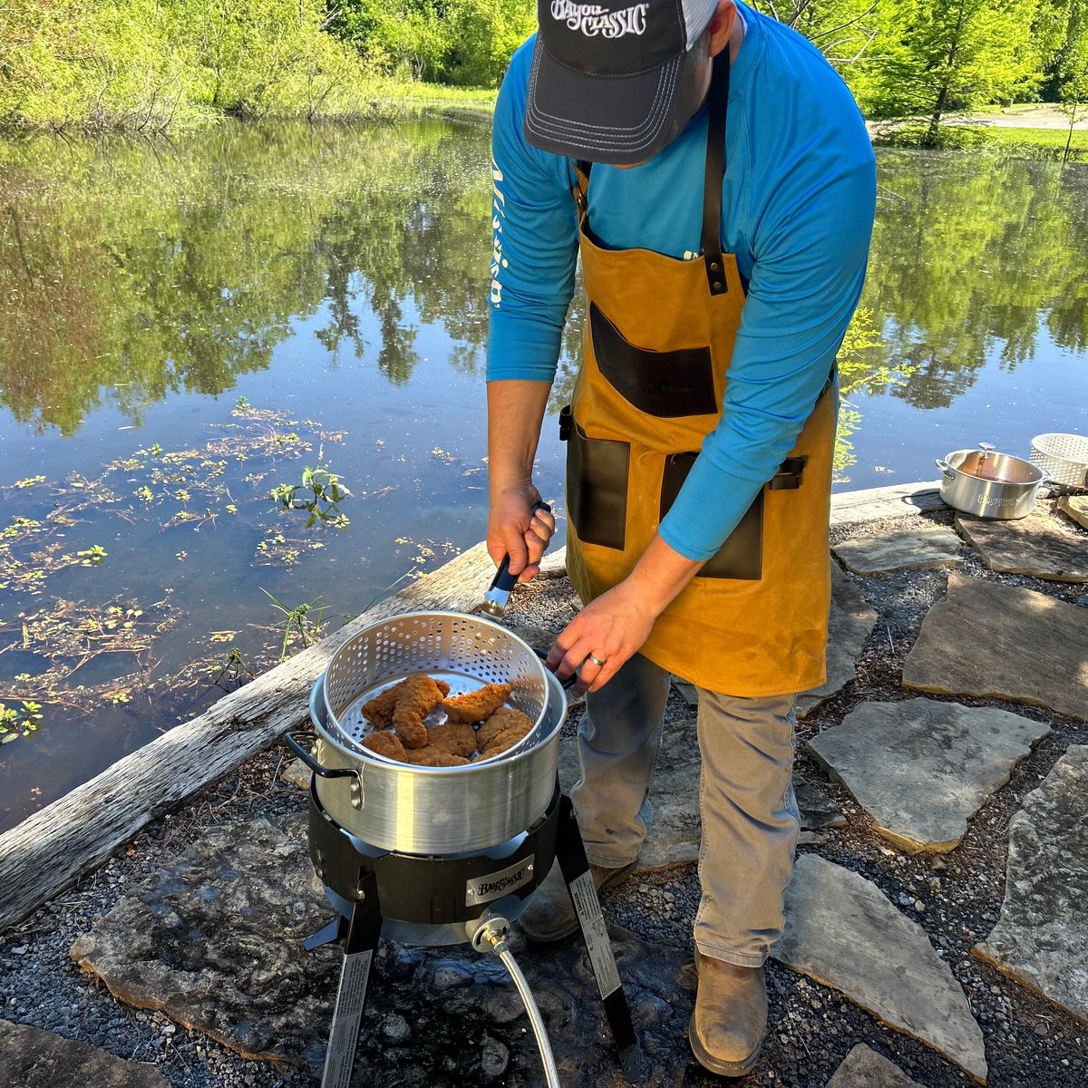 Person cooking outdoors by a lake using a fry pot and outdoor cooker.