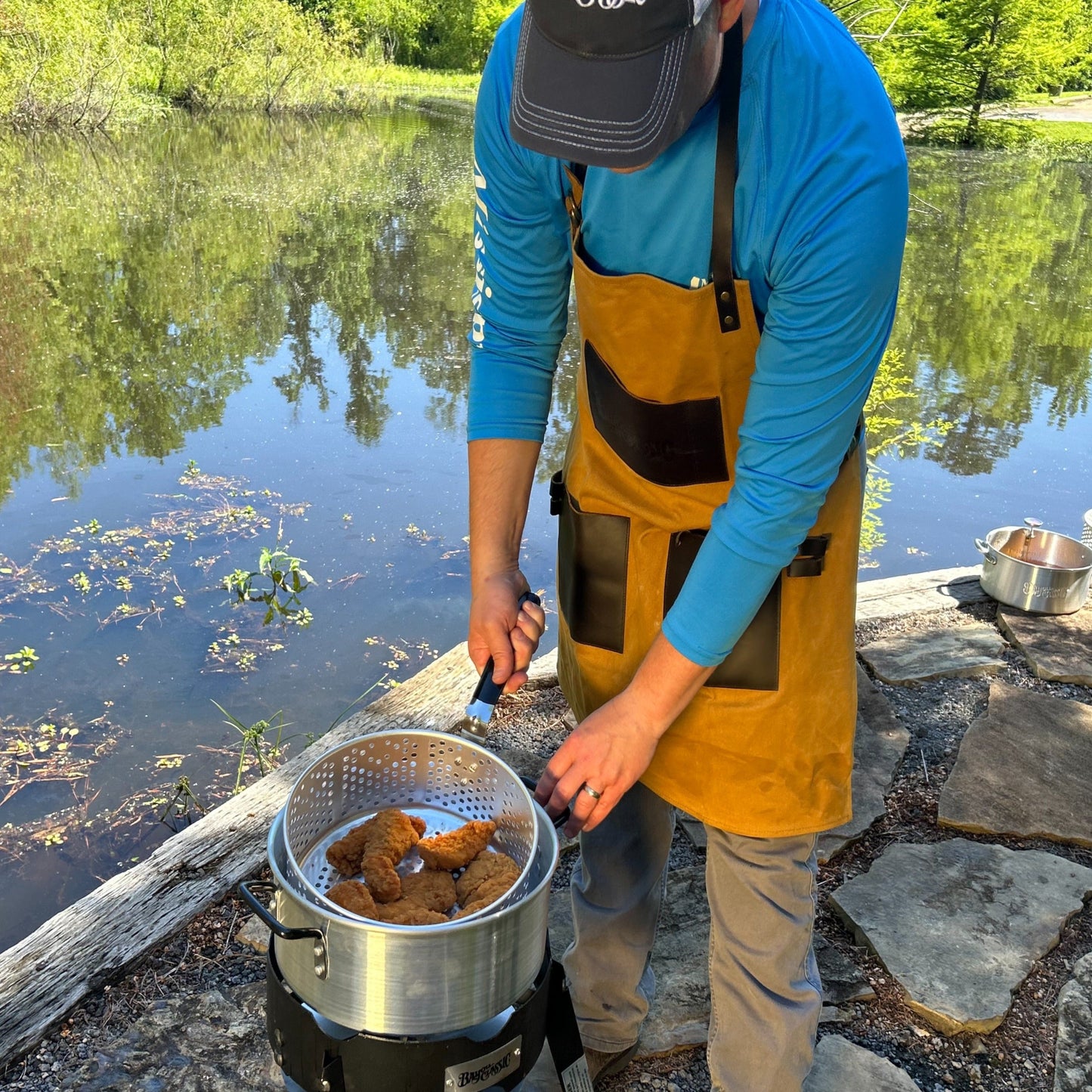 Person cooking outdoors by a lake, using a fry pot and outdoor cooker.