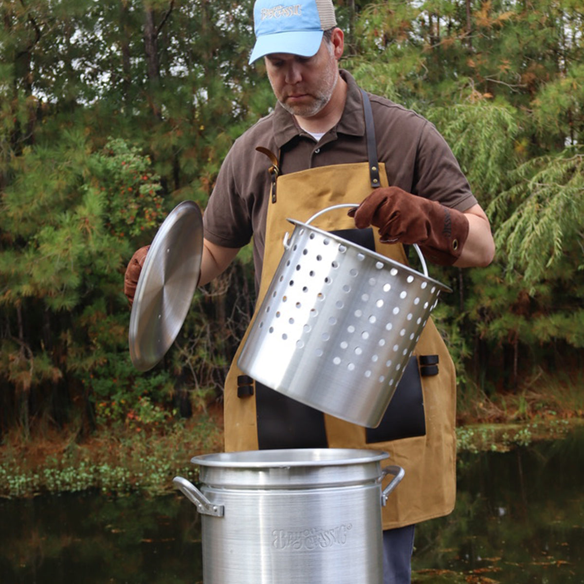 Person outside wearing an apron and brown fry gloves lifting a basket from a stockpot