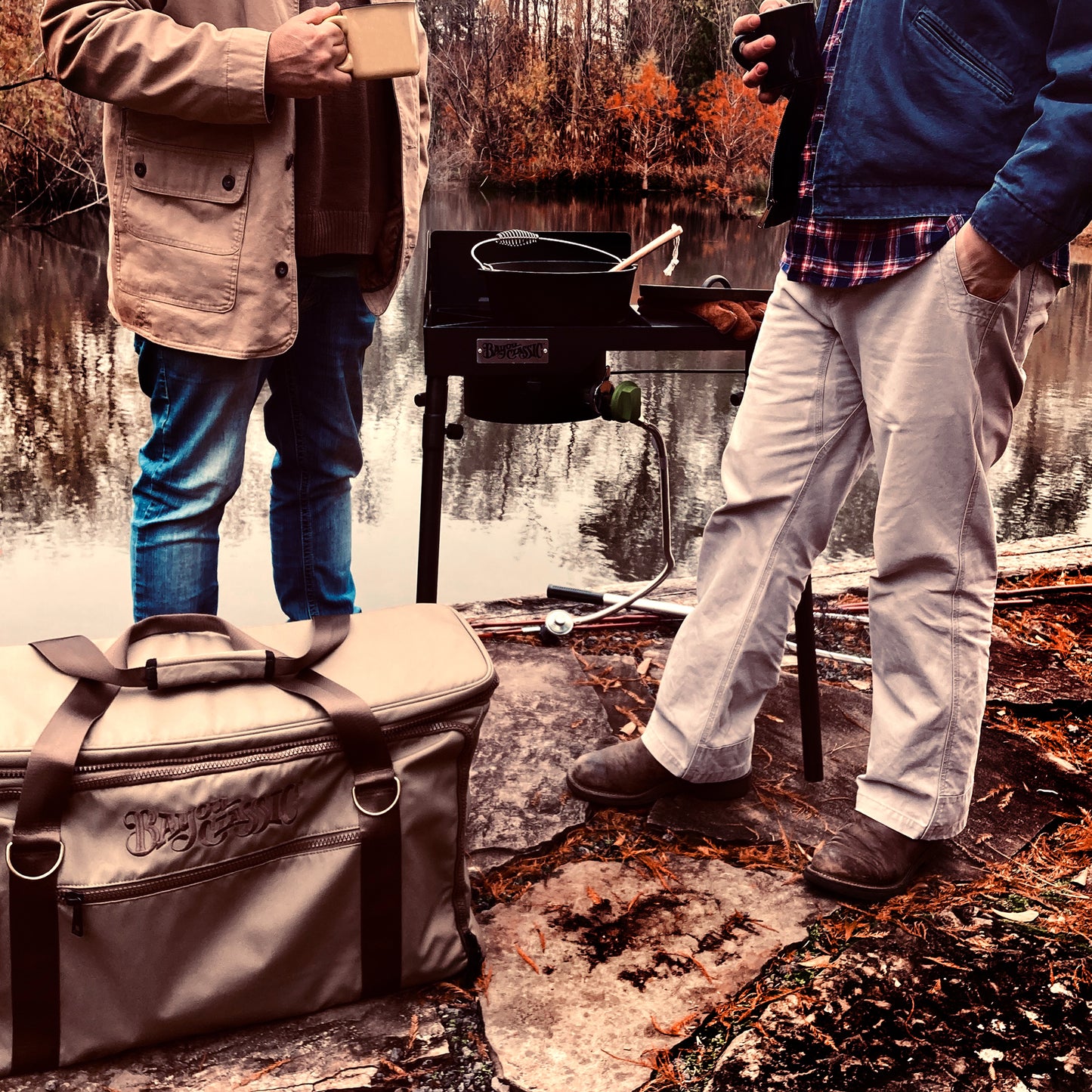 Two men outside by a body of water next to a cast iron cooker and gear bag