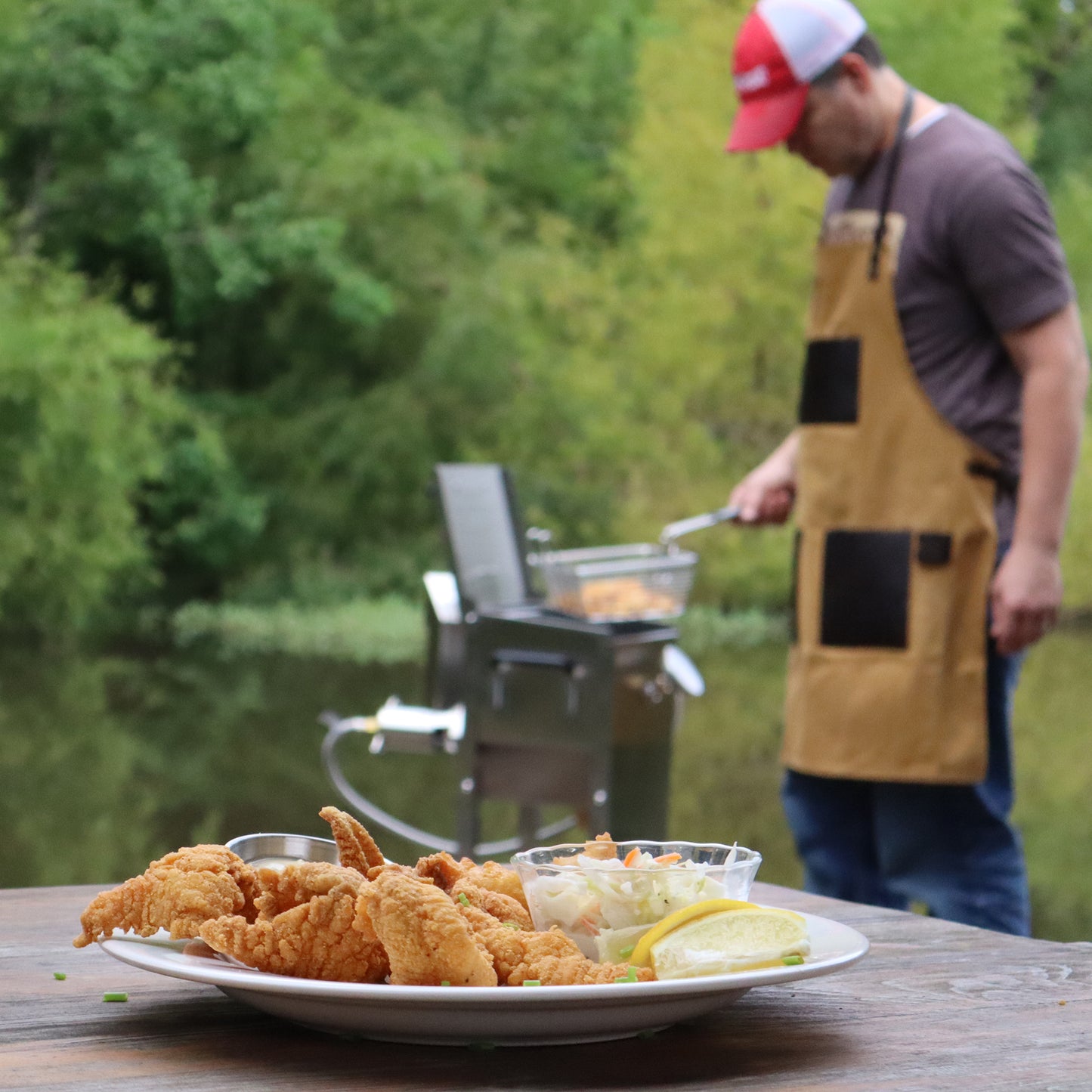 Man frying fish outside by a body of water. Plate of fish on a wood table.