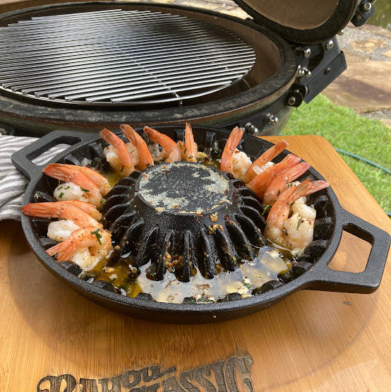 A black cast iron grill pan filled with shrimp and melted butter, sitting on a wooden outdoor table with a Bayou Classic grill in the background.