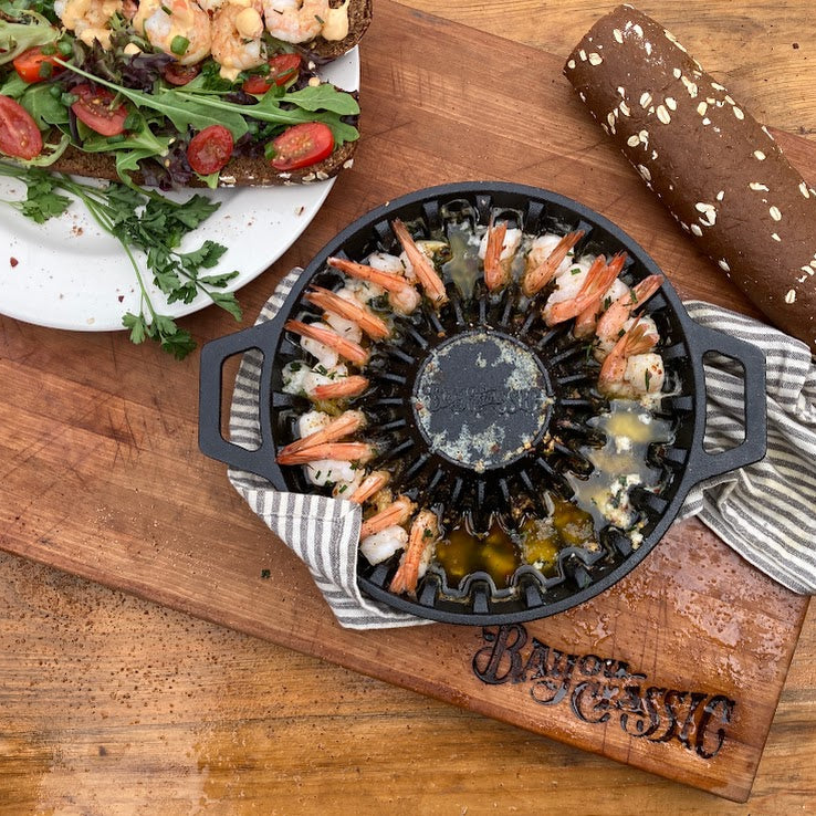 A round black cast-iron grill shrimp pan with cooked shrimp, placed on a wooden cutting board with a striped cloth underneath. A shrimp poboy and a bread roll are visible around it on a wooden table.