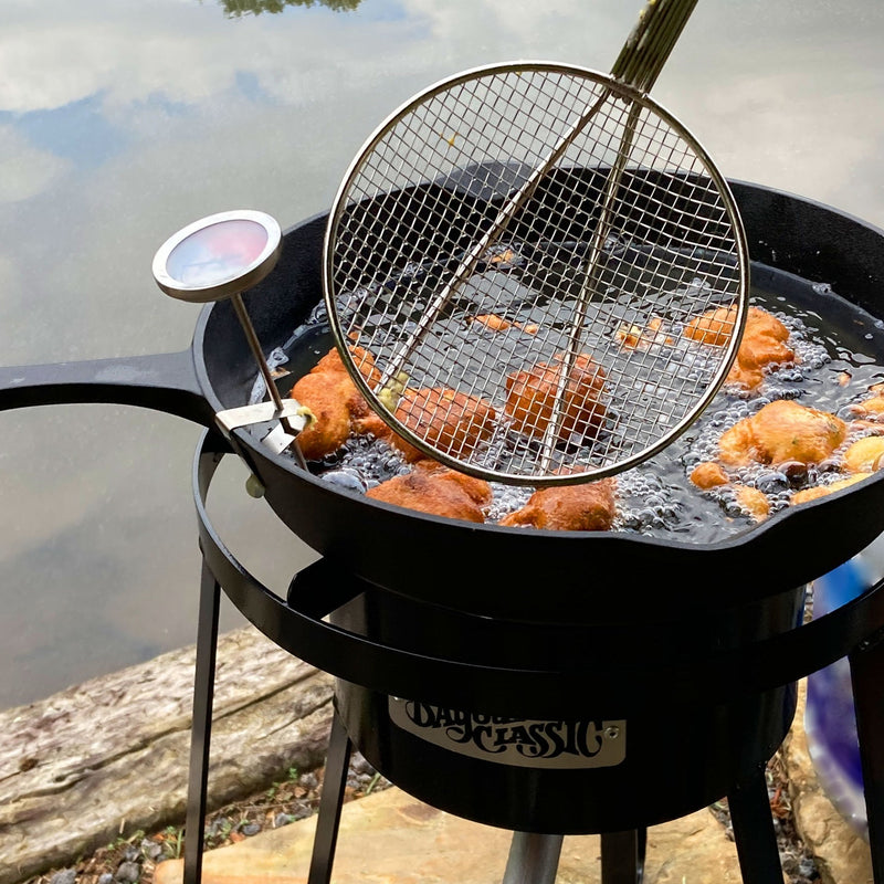 Cast iron skillet frying food over an outdoor cooker by a body of water