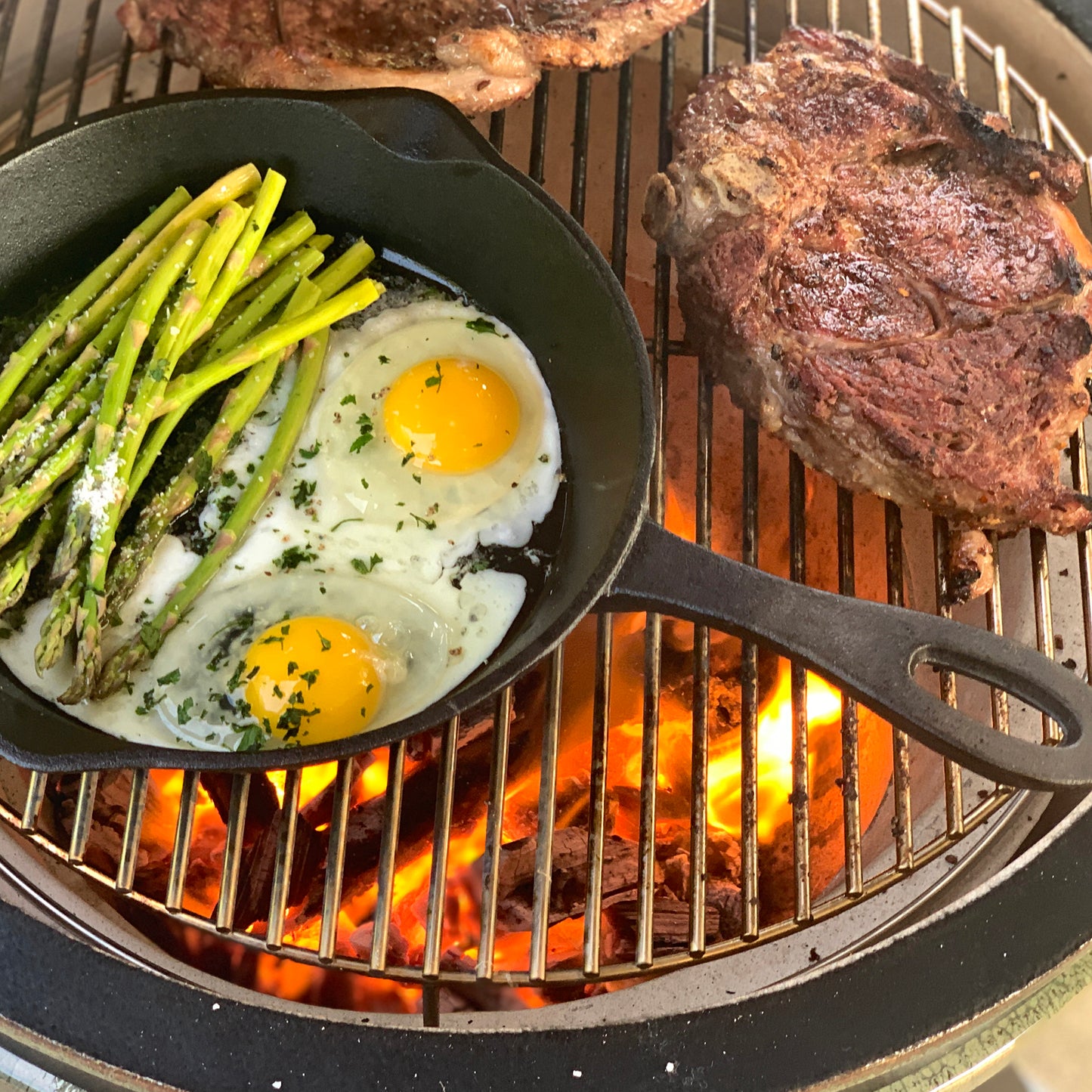 Cast Iron Skillet with fried eggs and asparagus on a grill next to two ribeye steaks