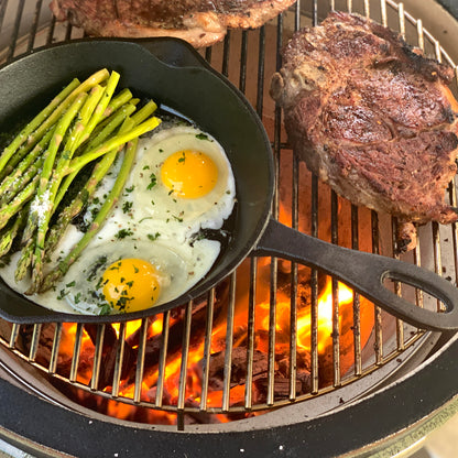 Cast Iron Skillet with fried eggs and asparagus on a grill next to two ribeye steaks
