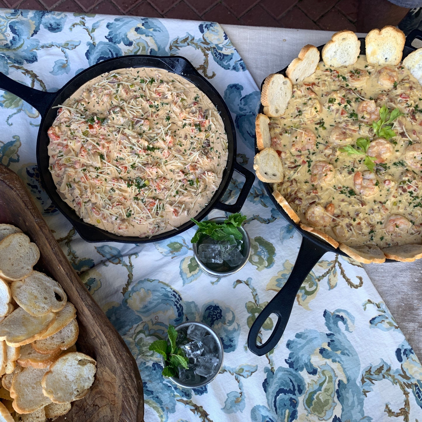 Two cast iron skillets of dip with bread slices on a patterned tablecloth.