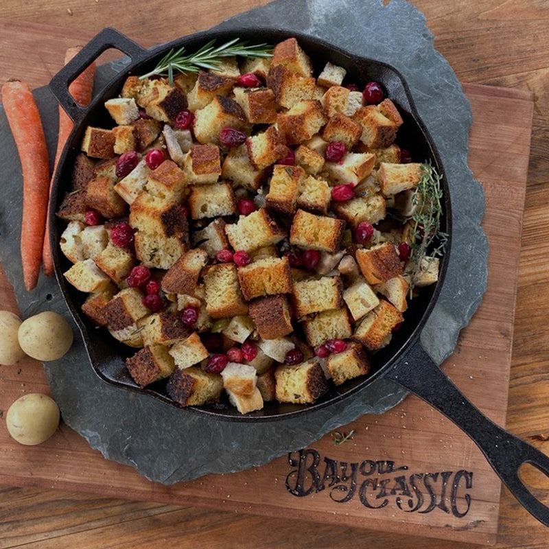 Skillet of stuffing with bread cubes and cranberries on a wooden board with 'Bayou Classic' branding.