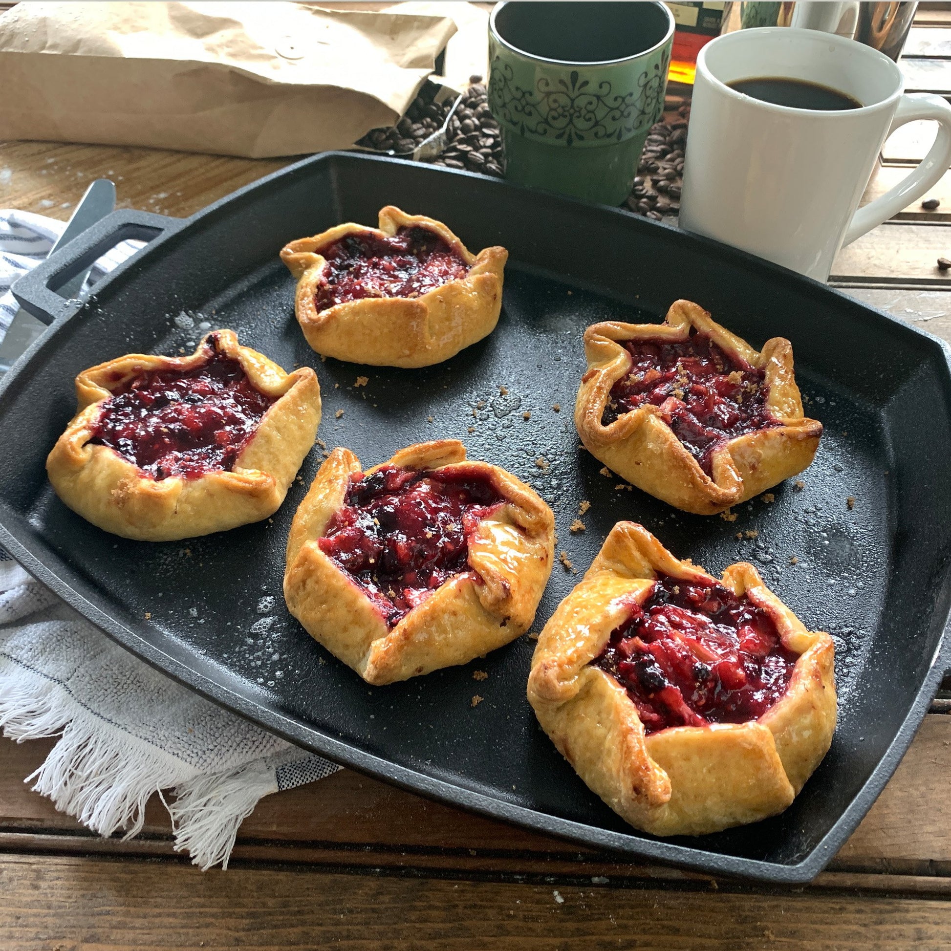 Pastry tarts with berry filling on a black pan with coffee and tea cups in the background.