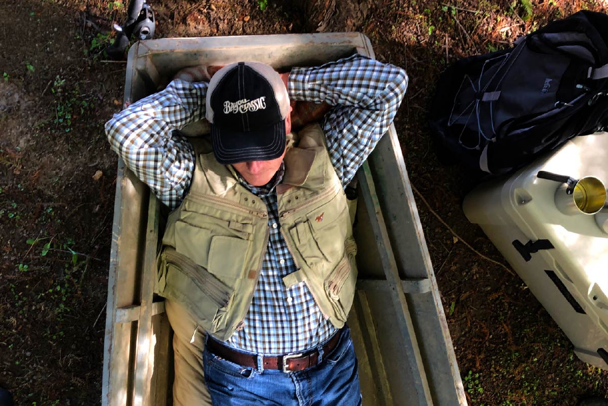 Man lying on a wooden cart with a cooler and backpack in an outdoor setting