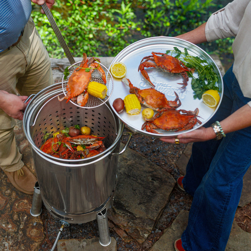 Outdoor cooking scene with a stainless steel pot and a large metal tray holding cooked crabs, corn, lemon slices, and greens, with two people partially visible handling the food near a pond.