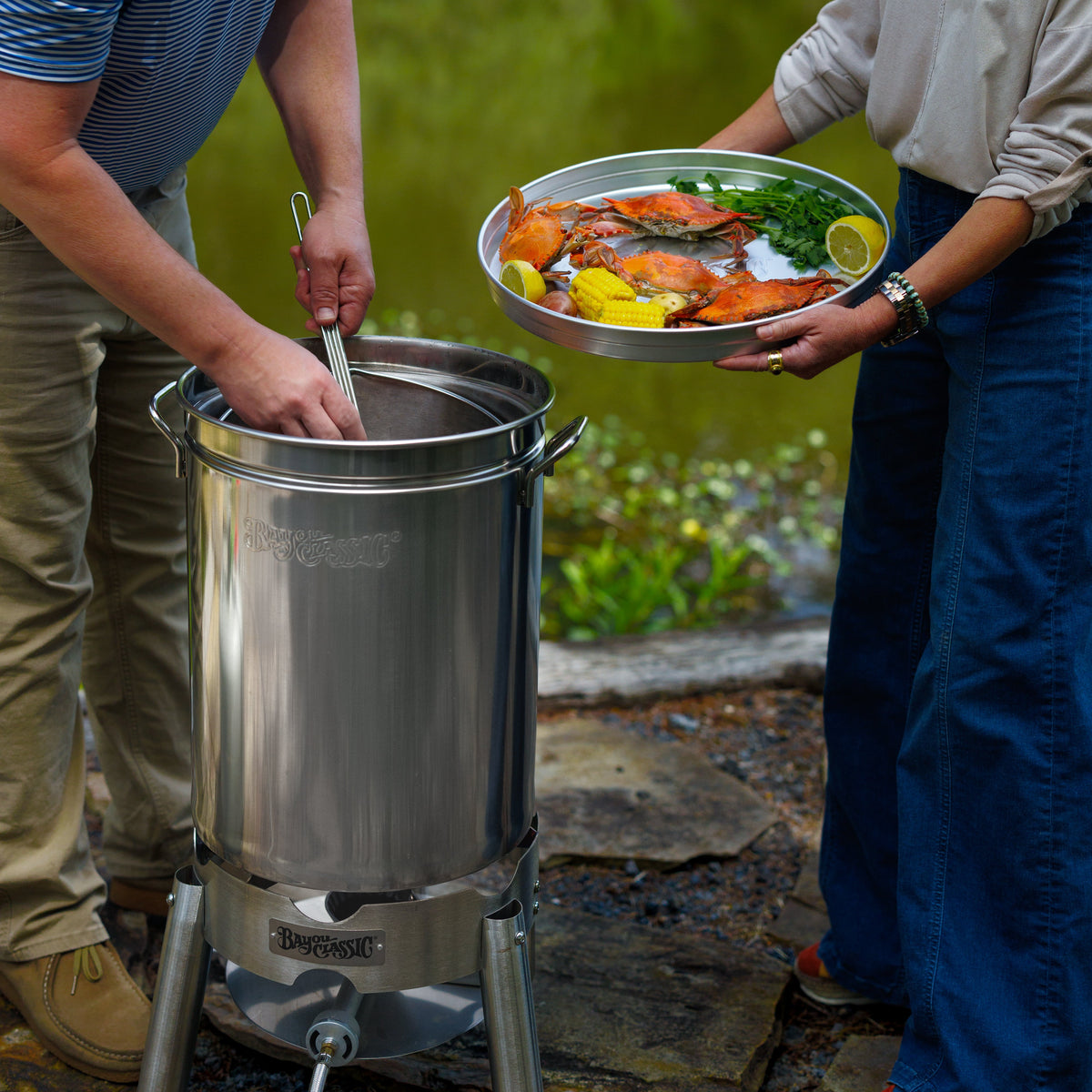 A stainless steel outdoor boiling pot and cooker setup with two people in a natural setting. One person serving crabs on a tray with lemons and seasoning in an outdoor setting.