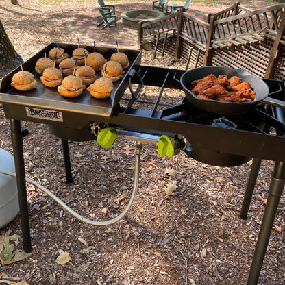 Double outdoor cooker with mini toasted burgers on a steel griddle over one burner and fried chicken in a cast iron skillet over the other burner