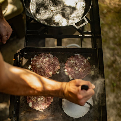 Person cooking burgers on a steel griddle on an outdoor cooker