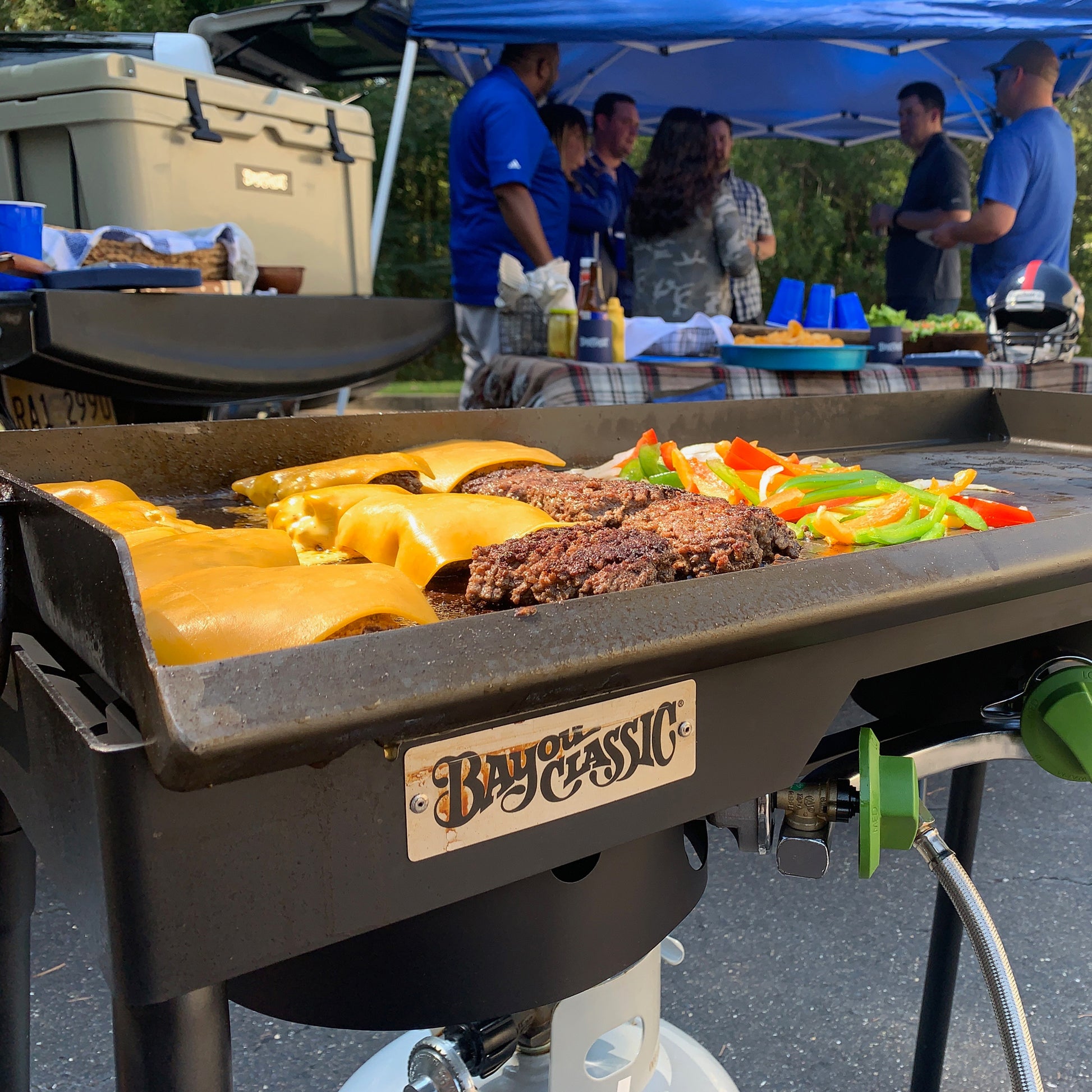 Steel griddle with cheeseburgers being cooked on a double outdoor cooker, people in the background under a tailgating tent