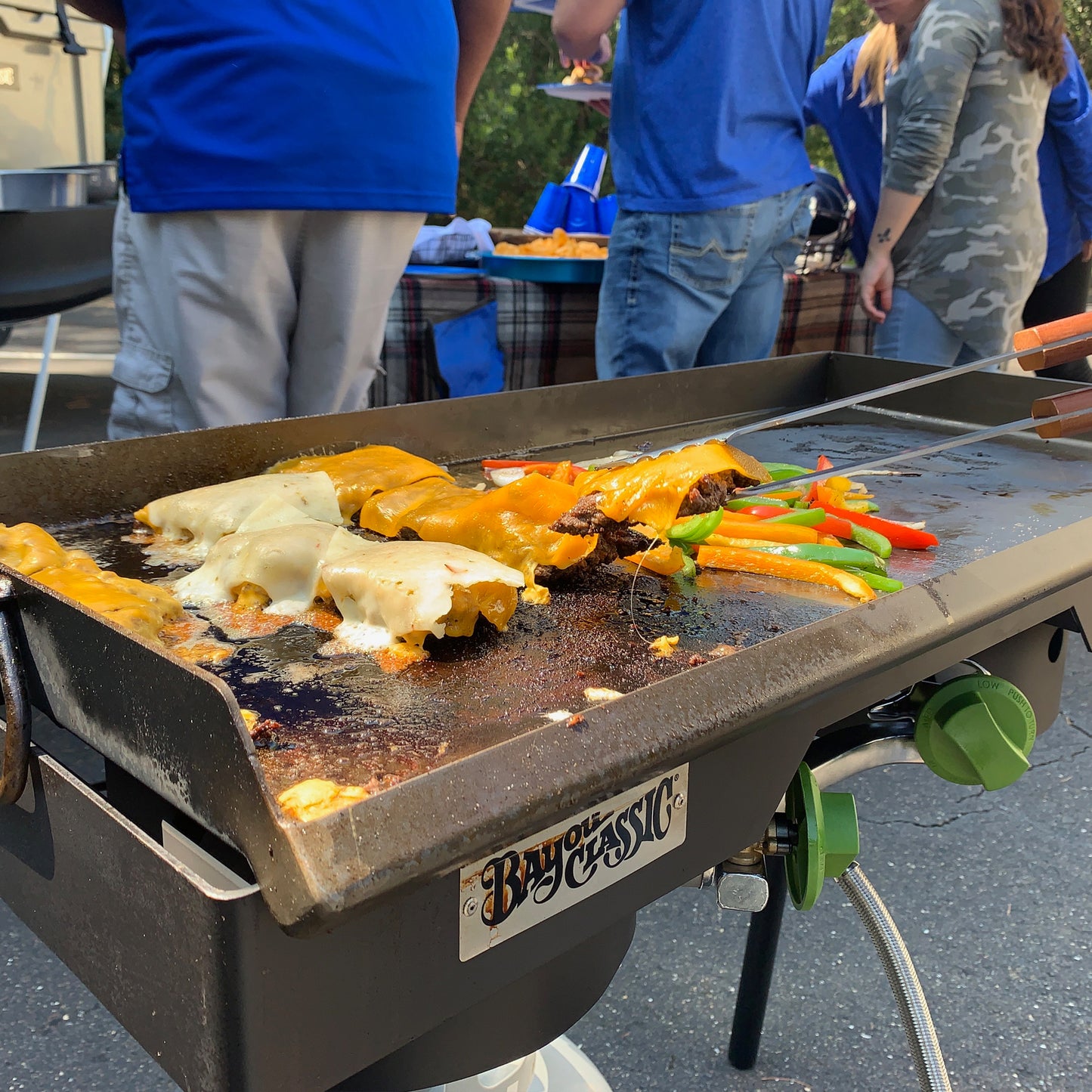 Steel griddle on a double outdoor cooker with cheeseburgers being cooked, people in the background