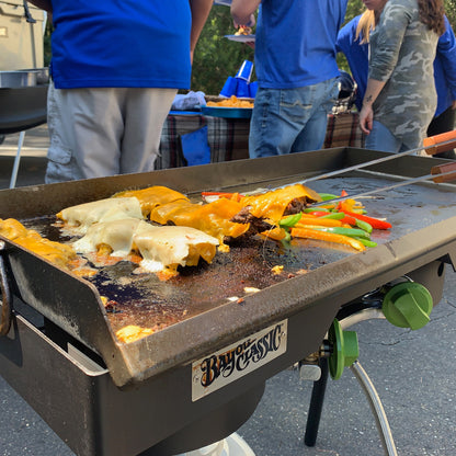 Steel griddle on a double outdoor cooker with cheeseburgers being cooked, people in the background