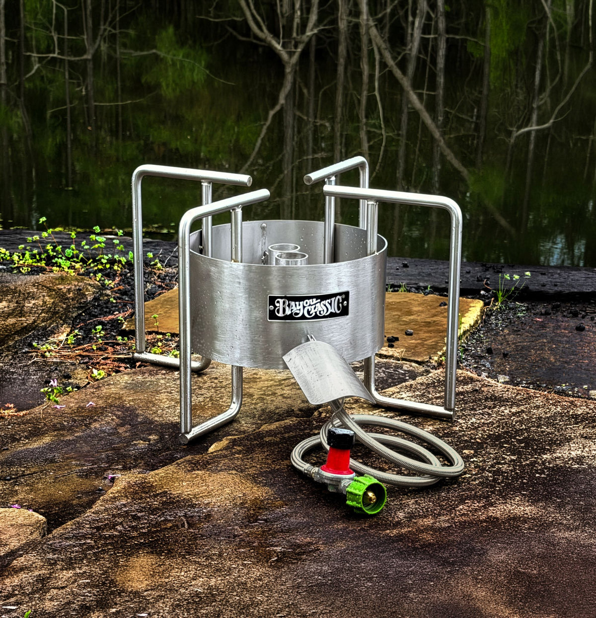 A stainless dual jet outdoor cooker, shown on a wet rock with a hose in the foreground and a pond in the background. 