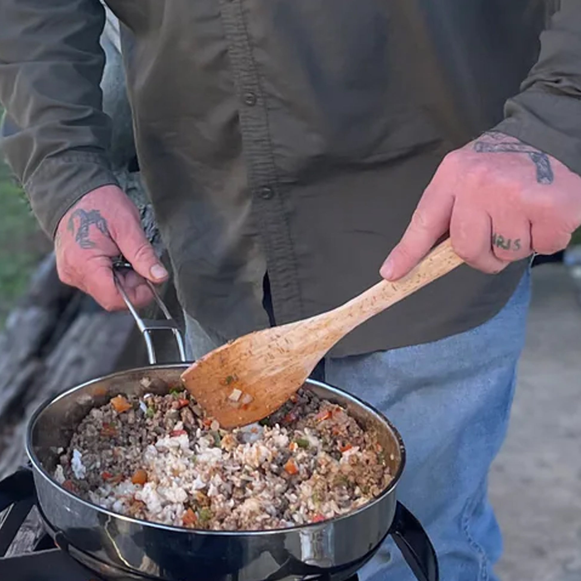 Person cooking outdoors with a stainless pan of food and wooden roux spoon