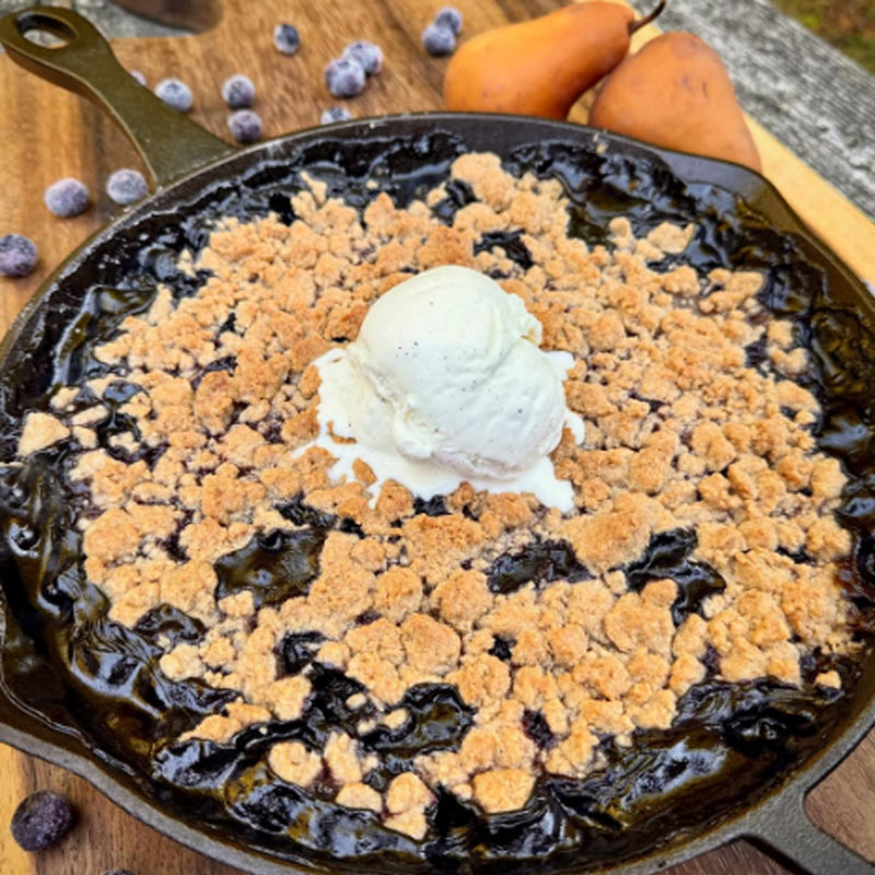 Blueberry cobbler with a scoop of ice cream in a cast iron skillet on a wooden surface.