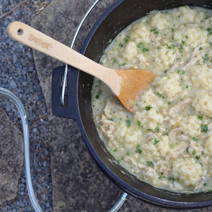 Chicken and dumplings being cooked in a cast iron dutch oven with a wooden roux spoon outside