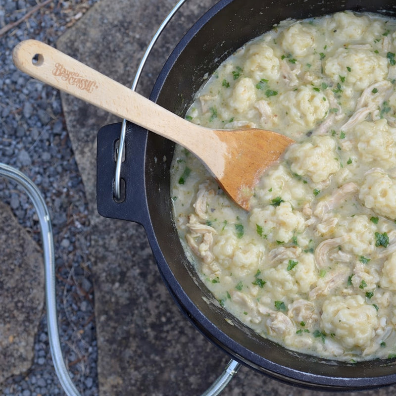 Chicken and dumplings being cooked in a cast iron dutch oven with a wooden roux spoon outside