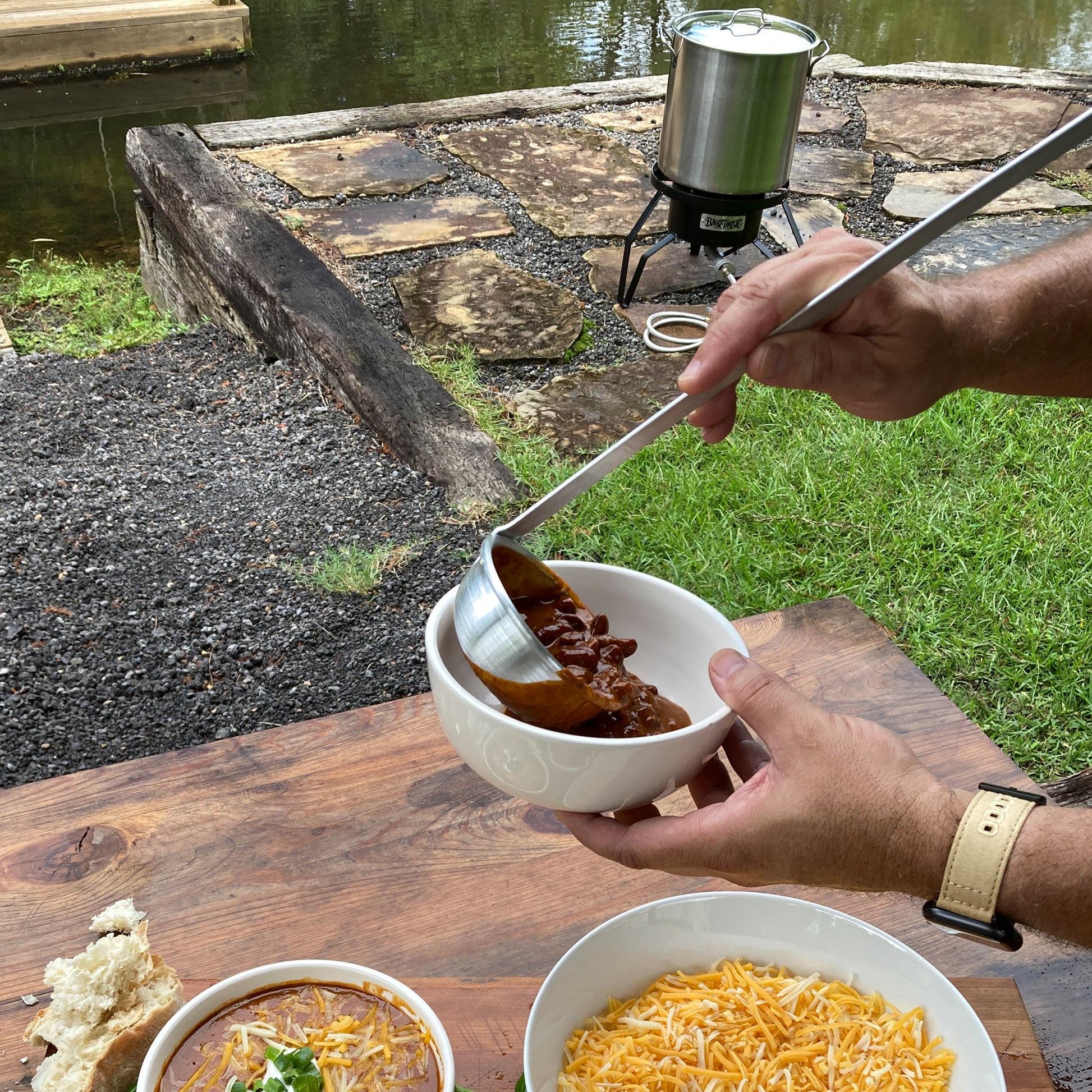 Person serving chili from a ladle into a bowl outdoors on a wooden table. Stockpot and outdoor cooker are in the background.
