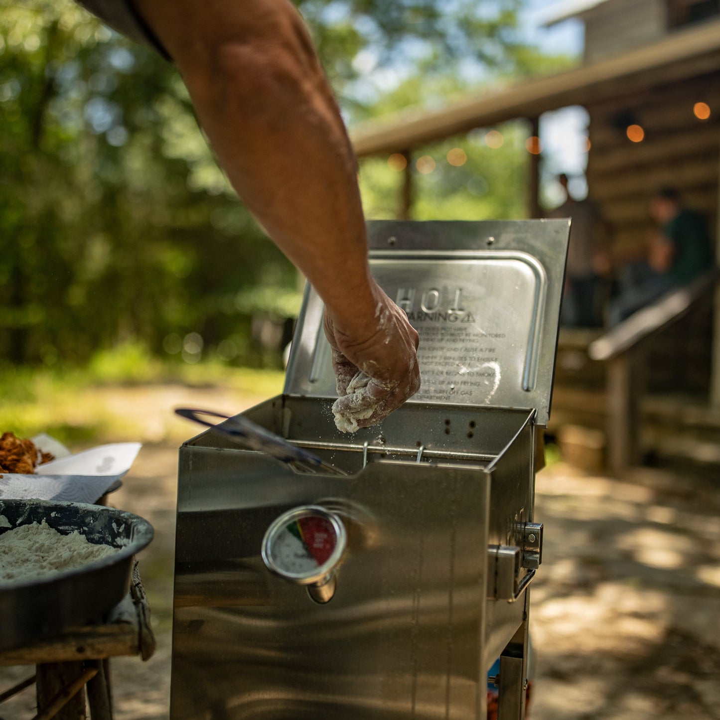 A stainless steel outdoor fryer with its lid open, shown in a backyard setting with a person's arm reaching into it. A log cabin and patio are visible in the blurred background.