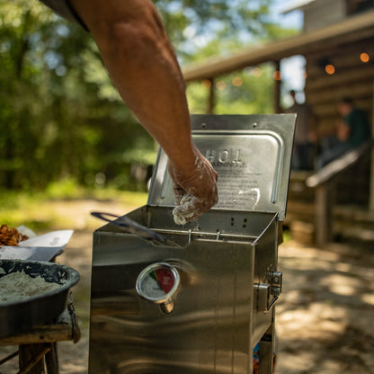 A stainless steel outdoor fryer with its lid open, shown in a backyard setting with a person's arm reaching into it. A log cabin and patio are visible in the blurred background.