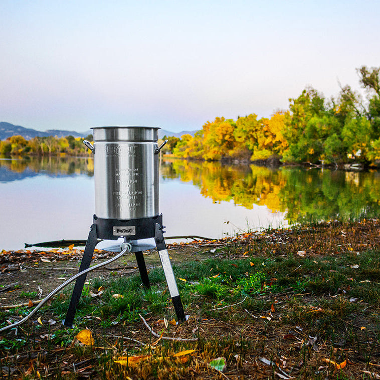 Stainless steel outdoor cooking pot on a tripod by a lake with trees in the background
