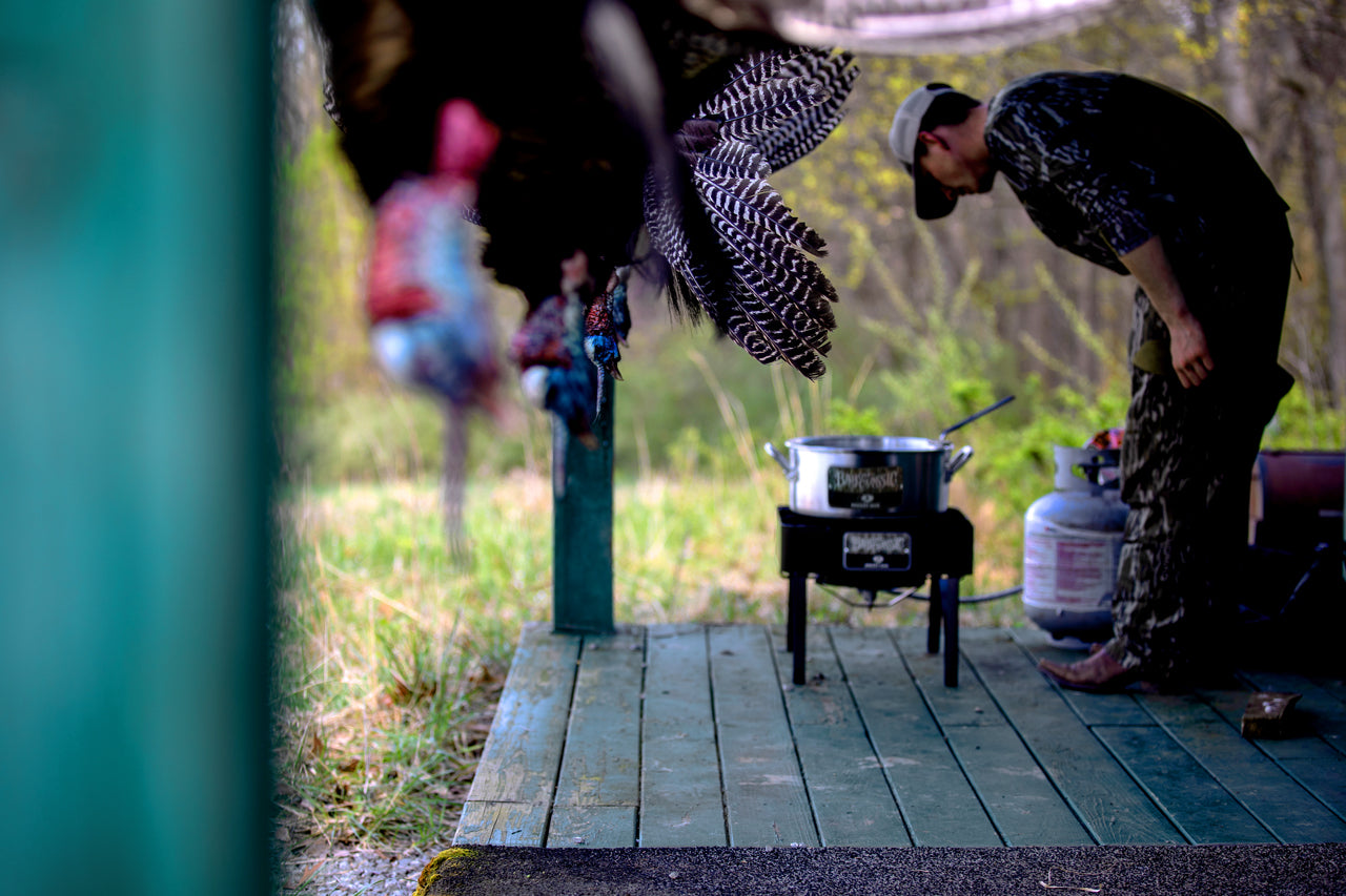Person cooking outdoors with a pot on a stove, surrounded by nature.
