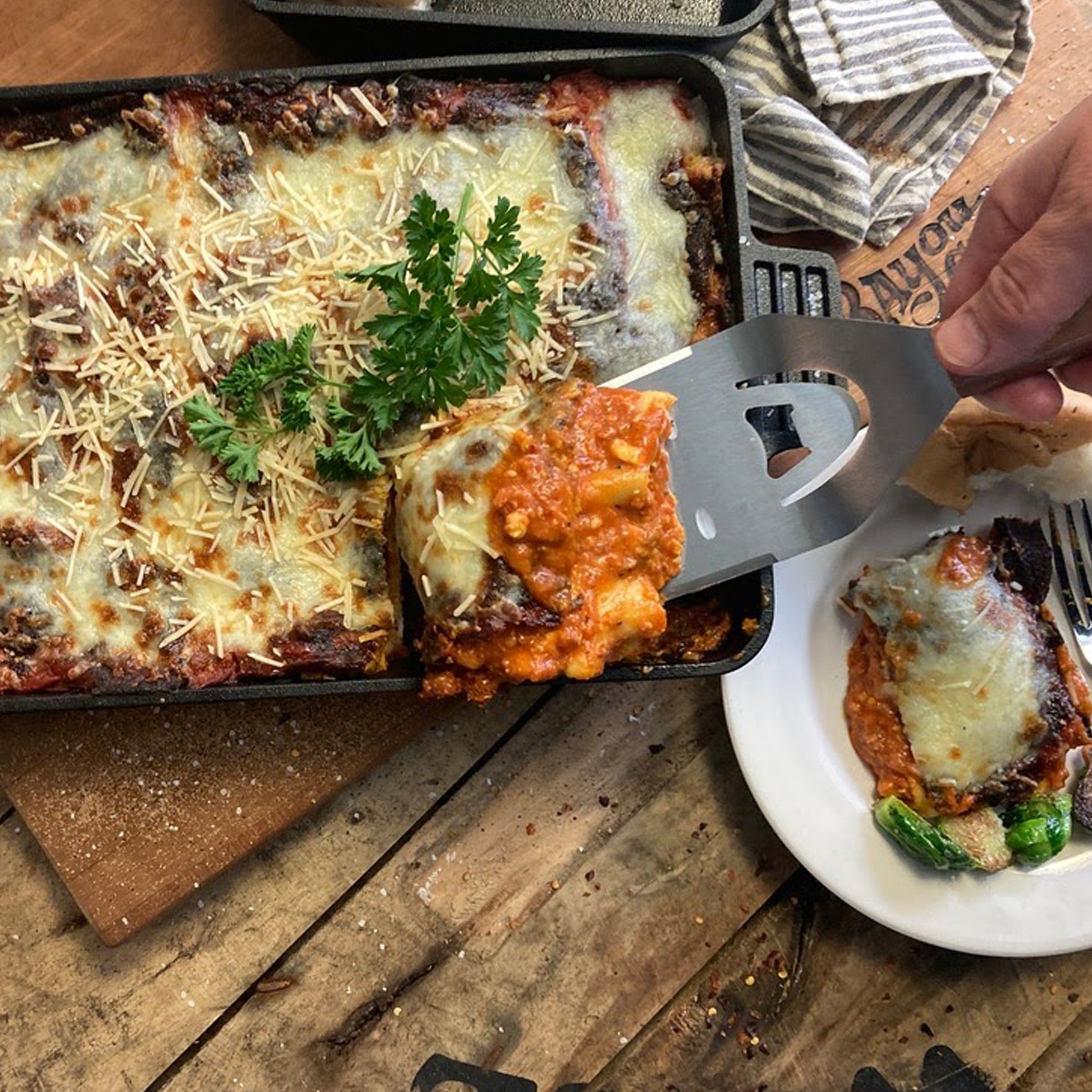 Baked lasagna in a cast iron baking pan being served with a spatula on a wooden table