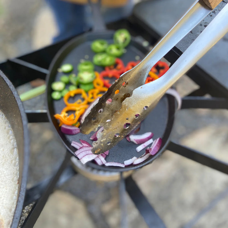 Using tongs to cook vegetables in a cast iron skillet on an outdoor cooker