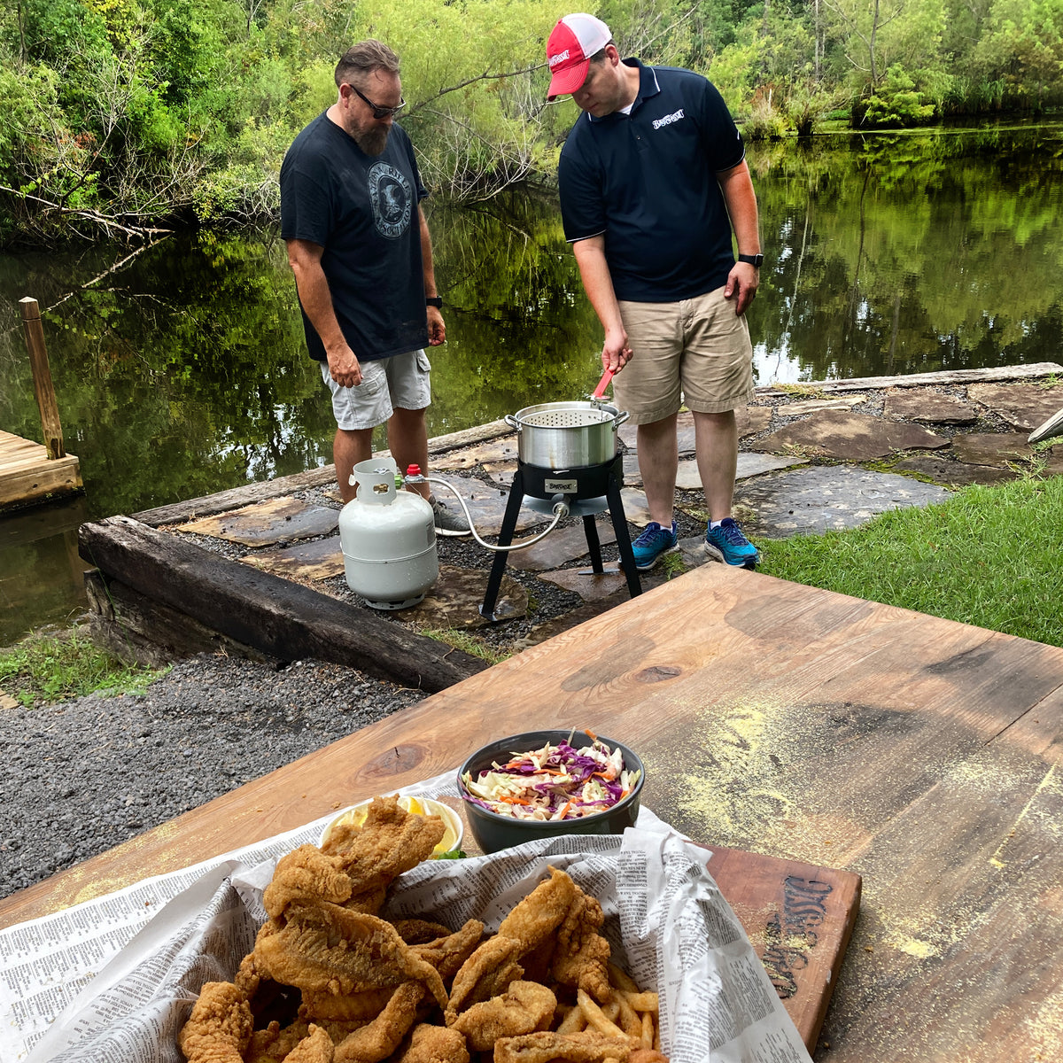 Two men frying fish outside by a body of water
