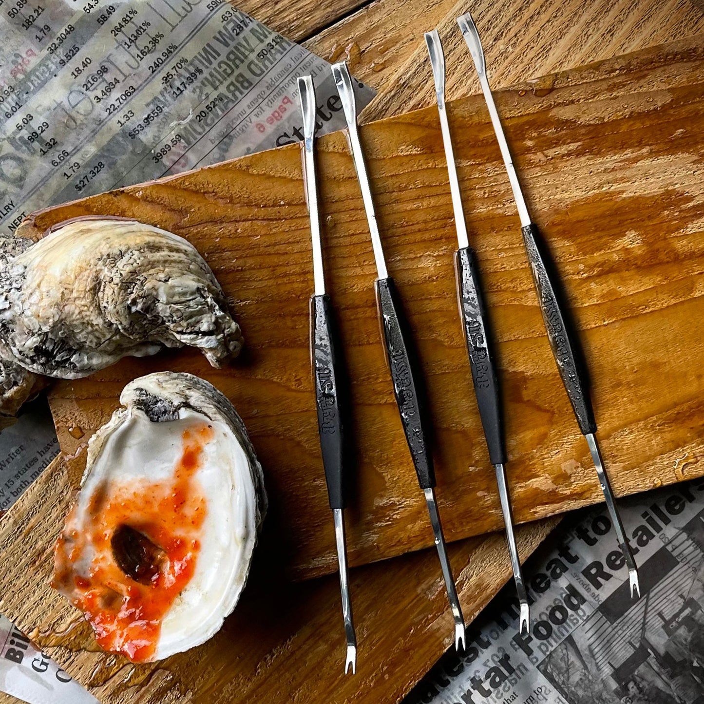A set of four stainless steel seafood forks with black handles, displayed alongside crab and lobster shells on a wooden surface.