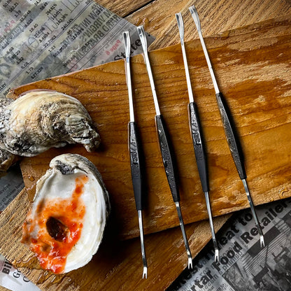 A set of four stainless steel seafood forks with black handles, displayed alongside crab and lobster shells on a wooden surface.