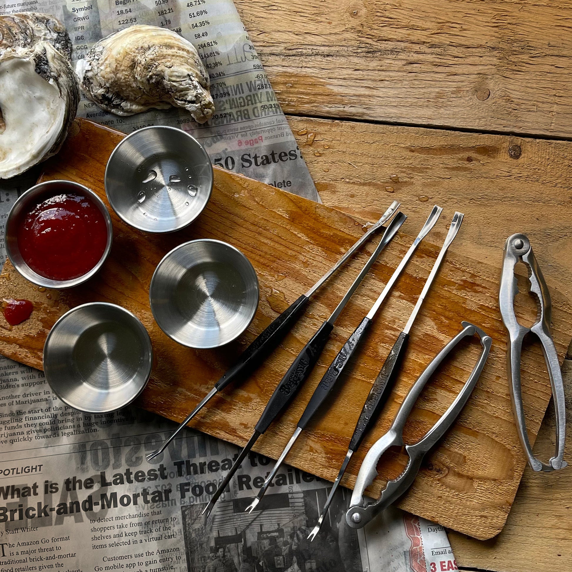 Set of seafood tools on a wooden surface with oyster shells and sauce in a ramekin