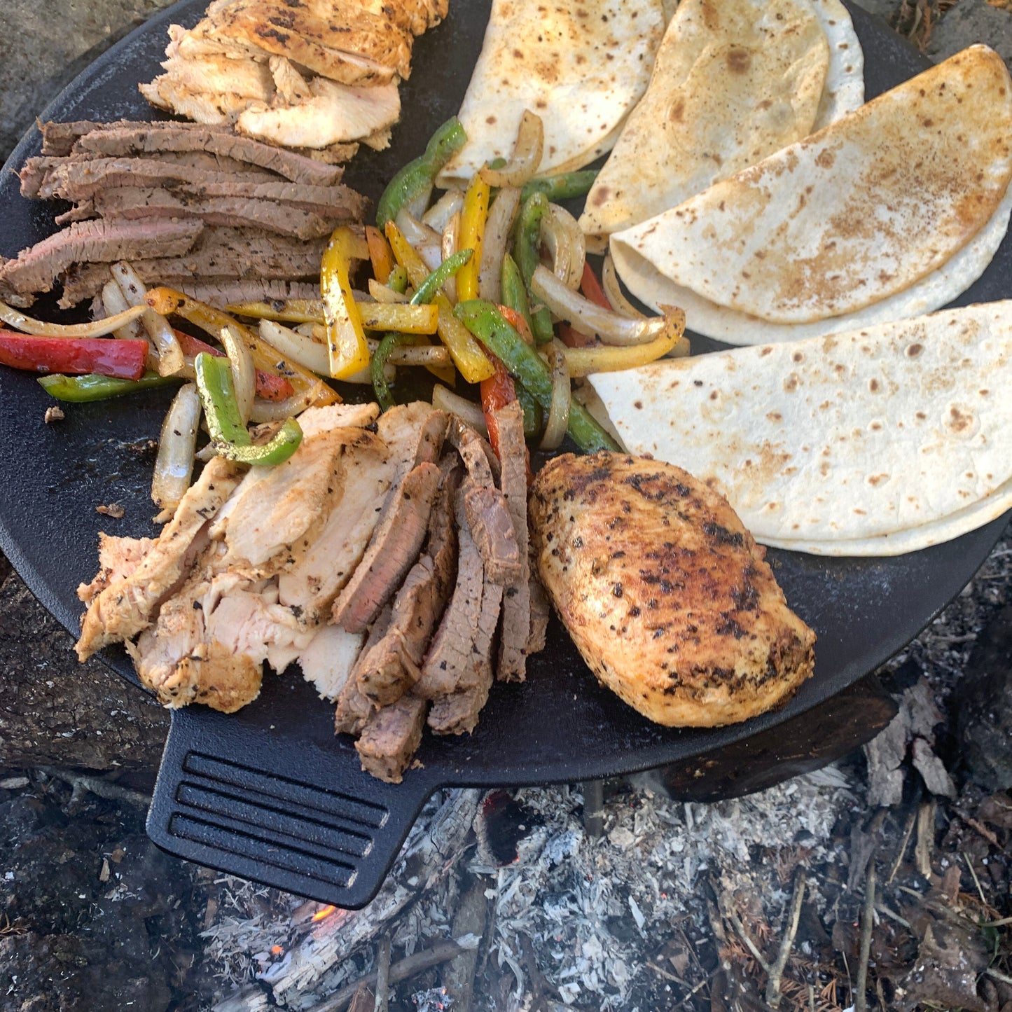 Cooked strips of steak and chicken with grilled peppers, onions, and fajita wraps on a cast iron discada over a campfire.