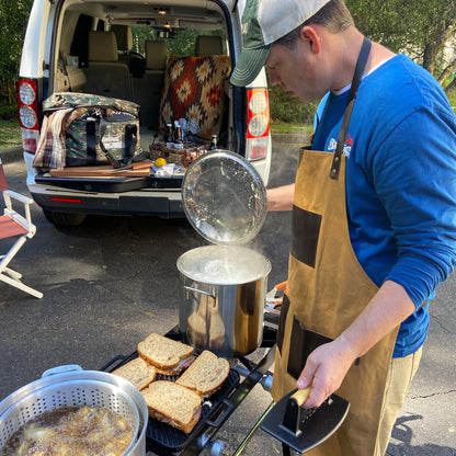 Person cooking at a car tailgate wearing a brown apron and using an outdoor cooker with a stockpot and griddle