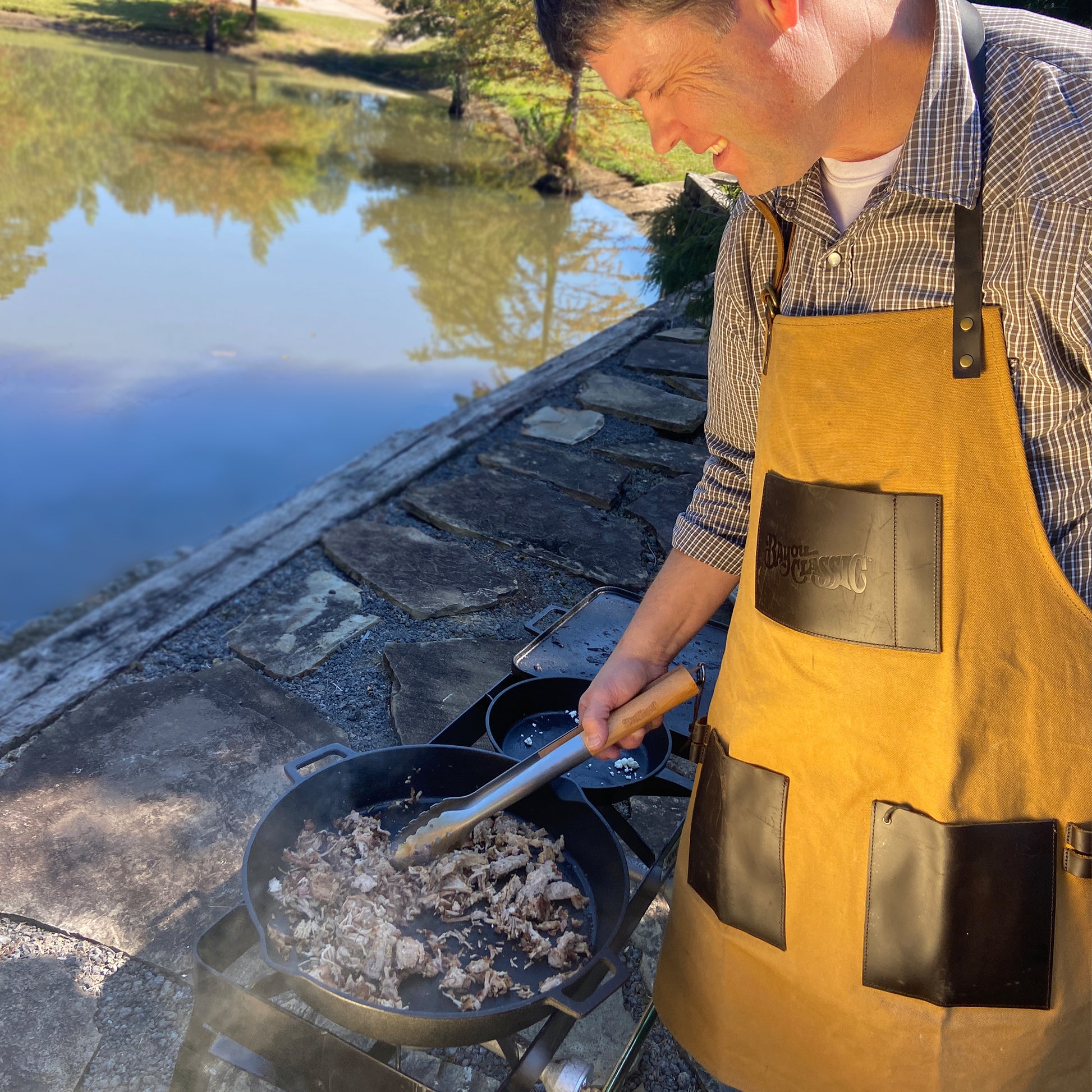 Man cooking outside wearing a brown apron and using a cast iron skillet over an outdoor patio stove